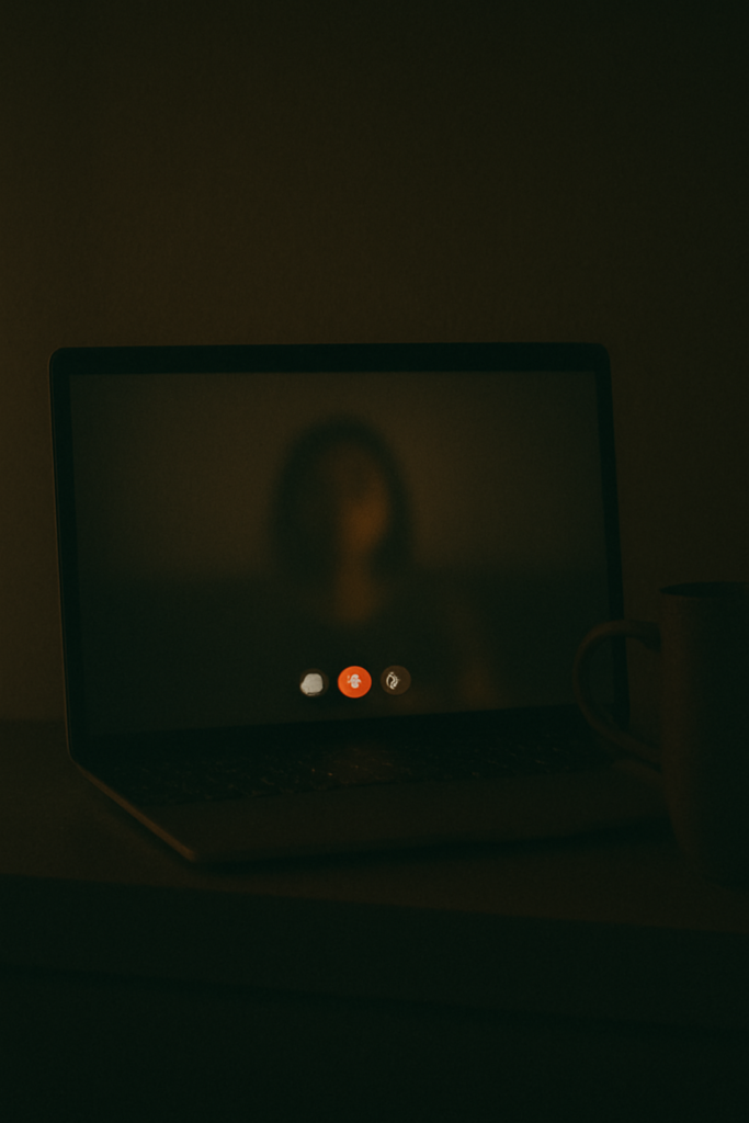 A laptop in a dark room displaying a blurry video call screen, with a faint reflection of a person and a ceramic mug nearby.